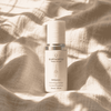 A white cosmetic serum bottle with a silver collar from quannessence Holistic Skincare, labeled 'QVitamin+ Replenishing Complex', styled on a linen surface. Serum contains advanced nano-encapsulated vitamin a, b3, c, and E.