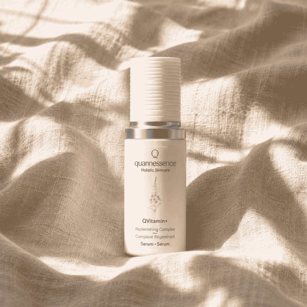 A white cosmetic serum bottle with a silver collar from quannessence Holistic Skincare, labeled 'QVitamin+ Replenishing Complex', styled on a linen surface. Serum contains advanced nano-encapsulated vitamin a, b3, c, and E.