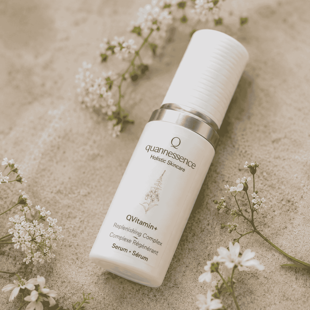 A white cosmetic serum bottle with a silver collar from quannessence Holistic Skincare, labeled 'QVitamin+ Replenishing Complex', styled on a textured beige surface with small white flowers. Serum contains advanced nano-encapsulated vitamin a, b3, c, and E. 