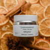 Jar of Quannessence Holistic Skincare QBreakout product on a background of dried oranges and cinnamon sticks.