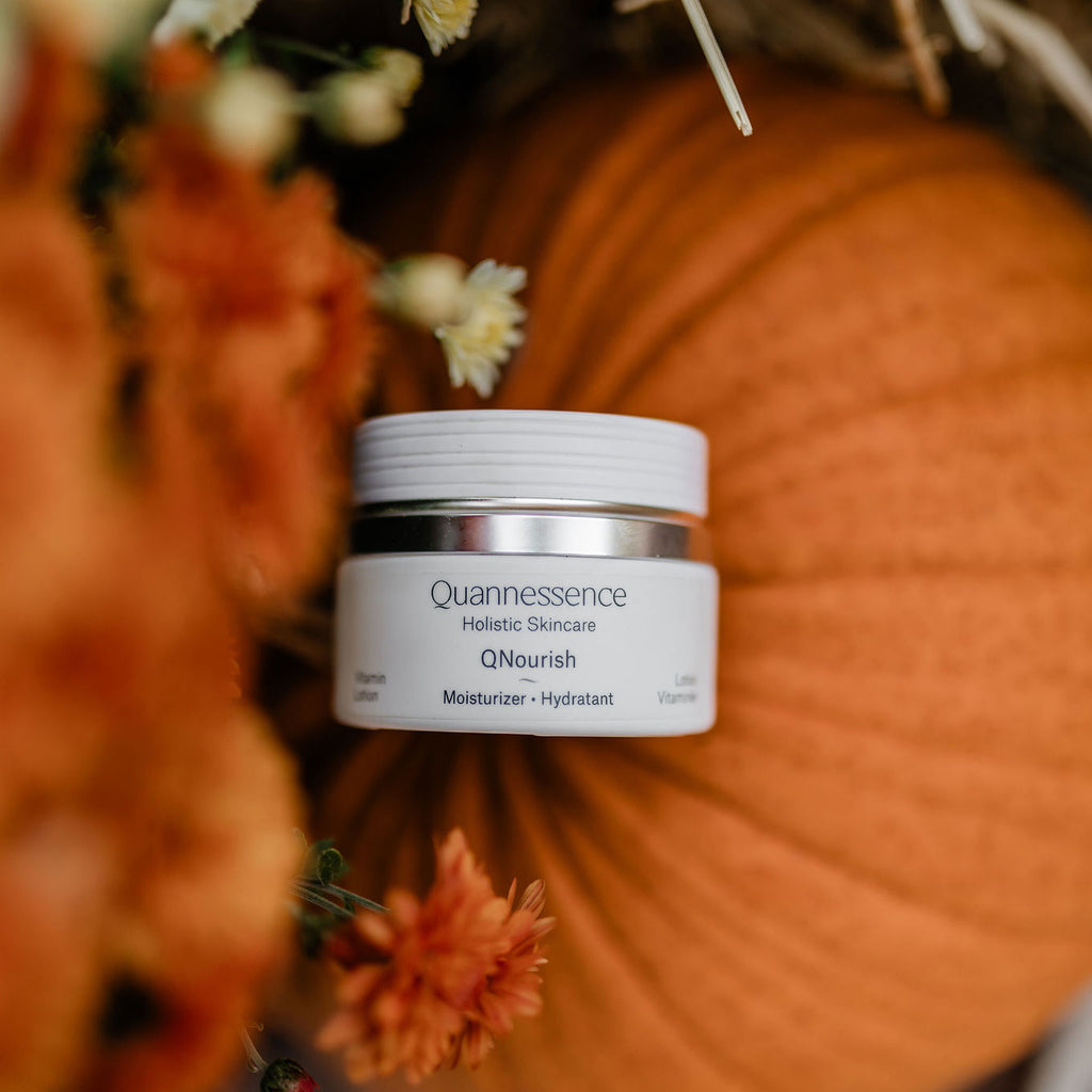 Skincare jar labeled 'Quannessence' on a pumpkin with flowers in the background. Quannessence Skincare, professional skincare, Holistic Beauty, Made in Canada, Naturally Sourced, Active ingredients, women-owned, Face, Moisturizer, Lotion, Cream, QNourish, uniQue encapsulated Vitamin Lotion, white jar, hand with cream
