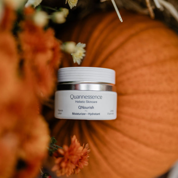 Skincare jar labeled 'Quannessence' on a pumpkin with flowers in the background. Quannessence Skincare, professional skincare, Holistic Beauty, Made in Canada, Naturally Sourced, Active ingredients, women-owned, Face, Moisturizer, Lotion, Cream, QNourish, uniQue encapsulated Vitamin Lotion, white jar, hand with cream