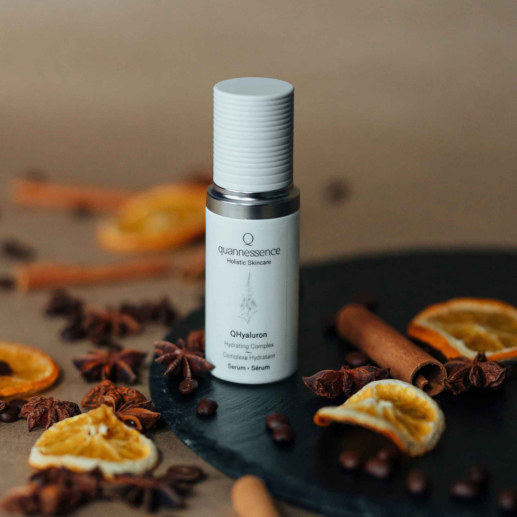 Skincare serum bottle labeled 'quannessence' on a dark surface with dried oranges and cinnamon sticks. Quannessence Skincare, professional skincare, Holistic Beauty, Made in Canada, Naturally Sourced, Active ingredients, women-owned, Face, Serum, QHYaluron, Hyaluronic (PUR) Hydrating Serum, white glass packaging with white lid & pump