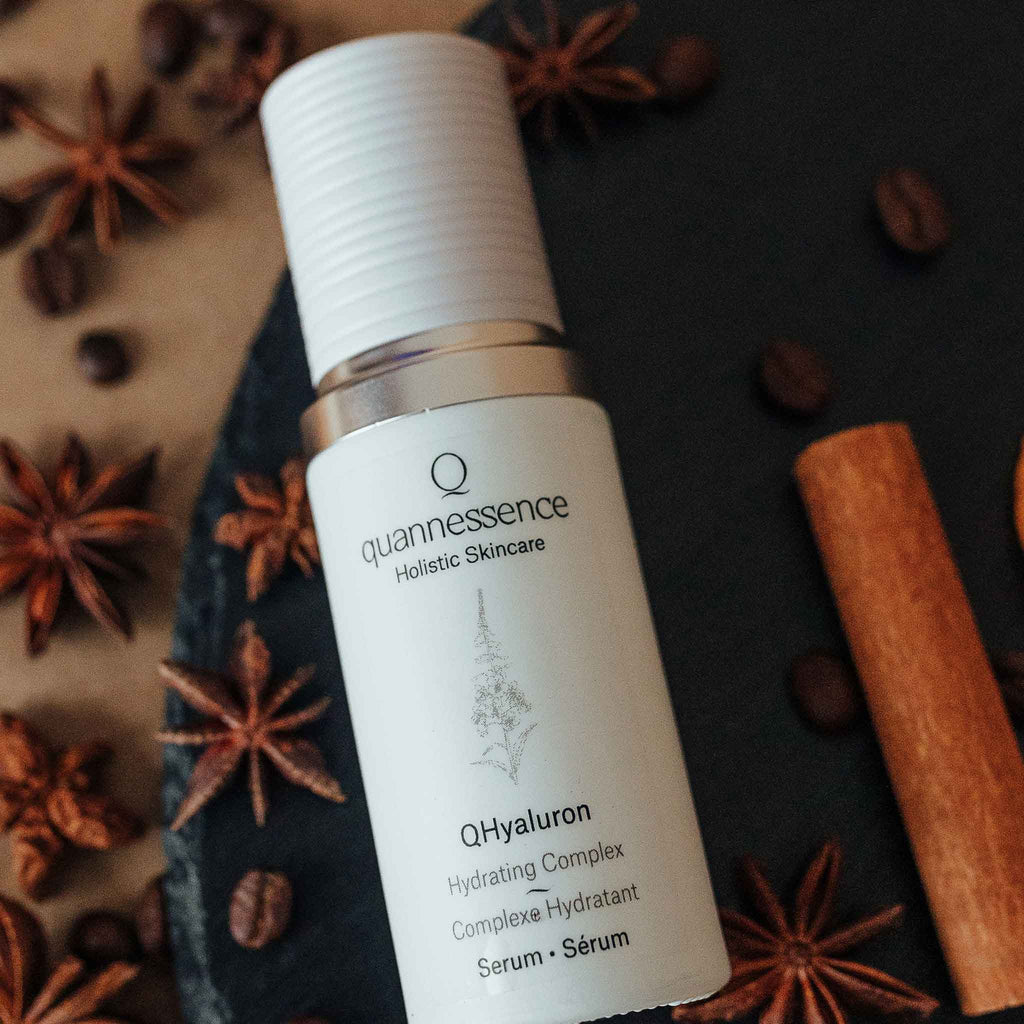 Skincare serum bottle labeled 'quannessence' on a dark surface with star anise and cinnamon sticks. Quannessence Skincare, professional skincare, Holistic Beauty, Made in Canada, Naturally Sourced, Active ingredients, women-owned, Face, Serum, QHYaluron, Hyaluronic (PUR) Hydrating Serum, white glass packaging with white lid & pump