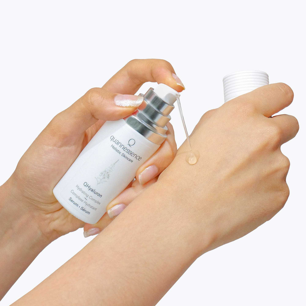 Person applying a droplet of a white serum bottle labeled 'quannessence' to their skin. Quannessence Skincare, professional skincare, Holistic Beauty, Made in Canada, Naturally Sourced, Active ingredients, women-owned, Face, Serum, QHYaluron, Hyaluronic (PUR) Hydrating Serum, white glass packaging with white lid & pump