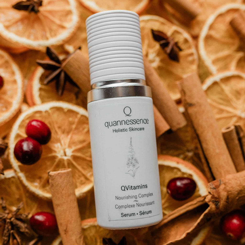 Quannessence Holistic Skincare serum bottle labeled 'quannessence' on a background of dried oranges, cinnamon sticks, and cranberries.